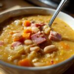 Close-up of a white bowl filled with thick, hearty bean soup featuring white beans, diced ham, and carrots.