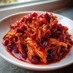 Close-up of shredded cranberry chicken coated in a rich, glossy cranberry sauce, served in a white bowl.