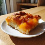 A square slice of warm peach cobbler with a thick, glistening, caramelized topping served on a small white plate.