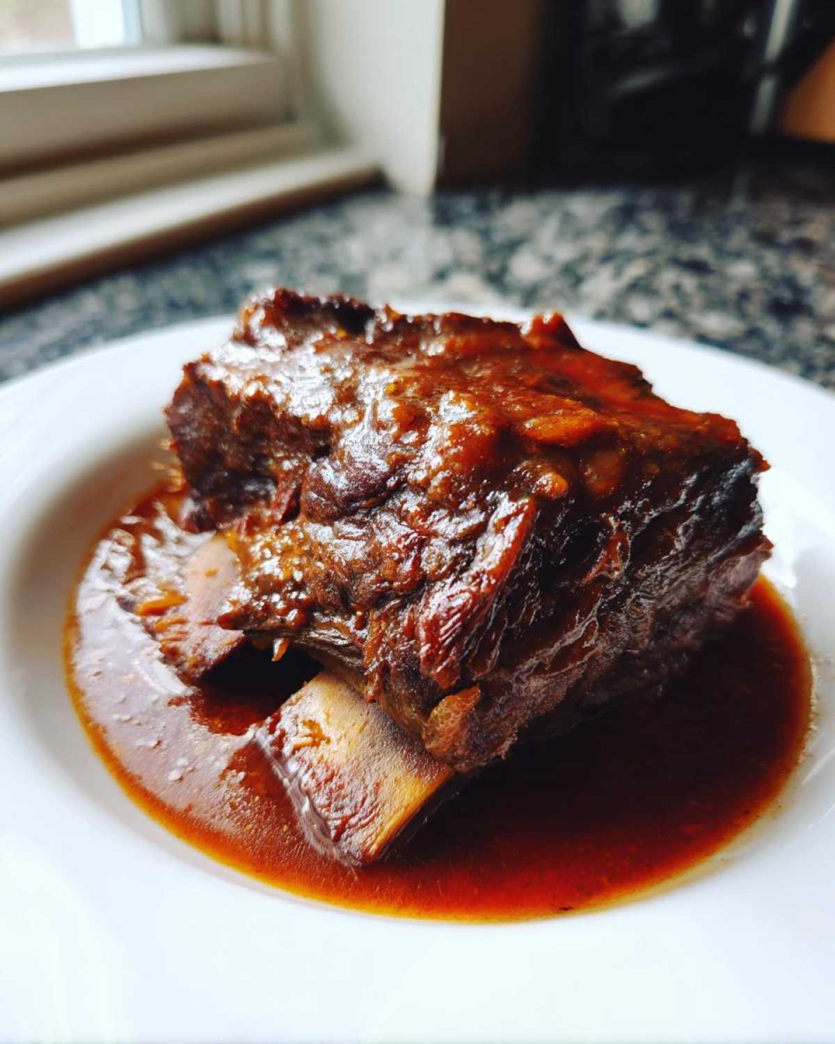 A single, rich, dark brown braised short ribs portion resting in a pool of savory sauce on a white plate.