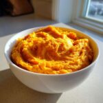 A close-up of fluffy, bright orange mashed sweet potatoes served in a white bowl.