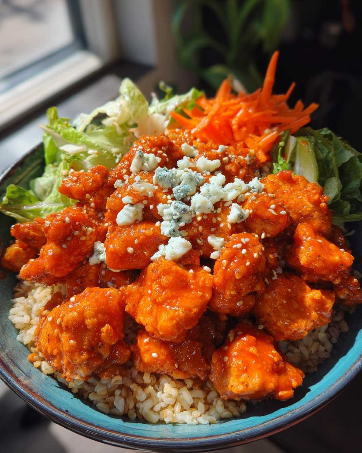 A vibrant buffalo chicken bowl featuring saucy chicken pieces over brown rice, topped with blue cheese crumbles and fresh vegetables.