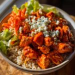 Close-up of a buffalo chicken bowl featuring saucy chicken pieces over brown rice, topped with blue cheese crumbles, lettuce, and shredded carrots.