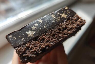 A close-up cross-section view of a rich, dark chocolate Capricorn cake slice featuring a black, glossy topping dusted with gold stars.