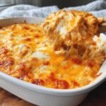 A wooden spoon scoops a cheesy, stringy portion of hot chicken enchilada dip from a white baking dish.