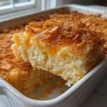 A serving of creamy, cheesy potato casserole being lifted from a white baking dish, topped with crispy cornflakes.
