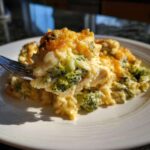 A fork lifts a creamy bite of chicken broccoli rice casserole topped with melted, browned cheese.