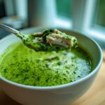 A spoonful of vibrant green chicken poblano soup, showing shredded chicken and herbs, lifted from a white bowl.