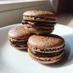 Close-up of three decadent chocolate macarons with glossy ganache filling stacked on a white plate.