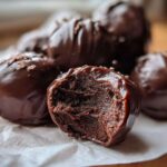 Close-up of rich, dark chocolate coated Oreo balls, with one bitten in half showing the dense, fudgy interior.