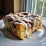 A gooey, frosted slice of cinnamon roll apple pie served on a white plate near a window.