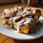 A close-up of a warm slice of cinnamon roll apple pie topped generously with white cream cheese icing.