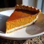 A close-up slice of classic pumpkin pie recipe showing a rich orange filling and a white cream layer on a white plate.