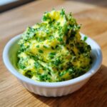 A close-up of fluffy, bright yellow cowboy butter dipping sauce mixed generously with chopped green herbs in a small white bowl.