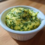 A close-up of bright yellow cowboy butter dipping sauce topped generously with chopped green herbs in a small white bowl.
