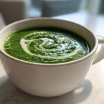 A vibrant green bowl of creamy spinach soup, garnished with a swirl of white cream.