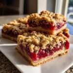 Three golden cranberry crumble bars stacked on a white plate, showing the thick cranberry filling.