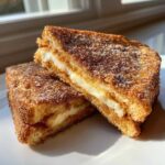 Two halves of golden brown cream cheese stuffed french toast dusted with cinnamon sugar, stacked on a white plate.