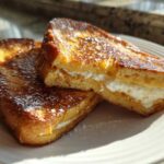 Two golden brown halves of cream cheese stuffed french toast dusted with powdered sugar on a white plate.