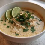 A close-up of a creamy keto white chicken chili, topped with shredded chicken, fresh cilantro, and two lime slices.