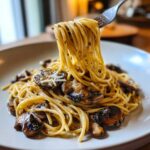 A fork lifting a generous twirl of creamy mushroom pasta from a white plate, showing sautéed mushrooms.