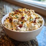 A white bowl filled with creamy Neiman Marcus dip, topped generously with bacon bits, shredded cheese, and chopped pecans.