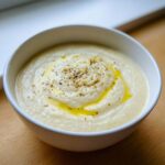 A close-up of a white bowl filled with creamy roasted cauliflower soup, drizzled with olive oil and sprinkled with pepper.