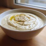 A close-up of creamy roasted cauliflower soup, swirled and topped with olive oil and herbs, in a white bowl.