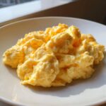 A close-up of perfectly cooked, creamy scrambled eggs piled high on a white plate, illuminated by sunlight.