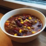 A close-up of a white bowl filled with rich crockpot cowboy soup, featuring ground meat, kidney beans, corn, and tomatoes.