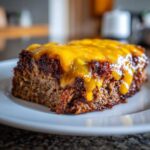 Close-up of a thick slice of crockpot french onion meatloaf topped with dripping, melted cheddar cheese on a white plate.
