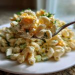 A fork lifts a creamy bite of deviled egg pasta salad with macaroni, celery, and crumbled egg yolk over a plate.