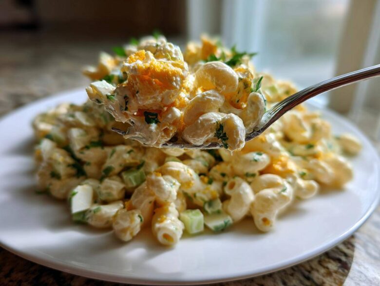 A fork lifts a creamy bite of deviled egg pasta salad with macaroni, celery, and crumbled egg yolk over a plate.