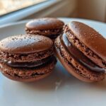 Three perfectly formed, rich brown chocolate macarons filled with glossy ganache, resting on a white plate.