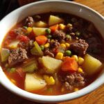 A close-up of a white bowl filled with rich hamburger soup featuring ground beef, potatoes, carrots, peas, and corn.