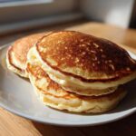 A stack of three perfectly golden brown almond flour pancakes resting on a white plate near a window.