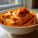 A white bowl filled with vibrant, fluffy mashed sweet potatoes, set near a window.