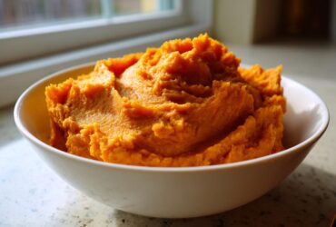 A white bowl filled with vibrant, fluffy mashed sweet potatoes, set near a window.