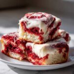A stack of three fluffy red velvet cinnamon rolls generously topped with white cream cheese icing.
