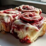 Three fluffy red velvet cinnamon rolls drizzled generously with white cream cheese icing on a white plate.