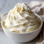 A close-up of light, fluffy vanilla buttercream frosting piped high in a white bowl.