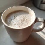 Close-up of a mug filled with creamy, foamy white hot chocolate, catching warm sunlight.