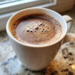 Close-up of a steaming mug filled with rich, frothy hot cocoa mix, set on a granite countertop.