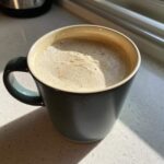 A close-up of a mug filled with creamy, frothy white hot chocolate, sitting on a light countertop in the sunlight.
