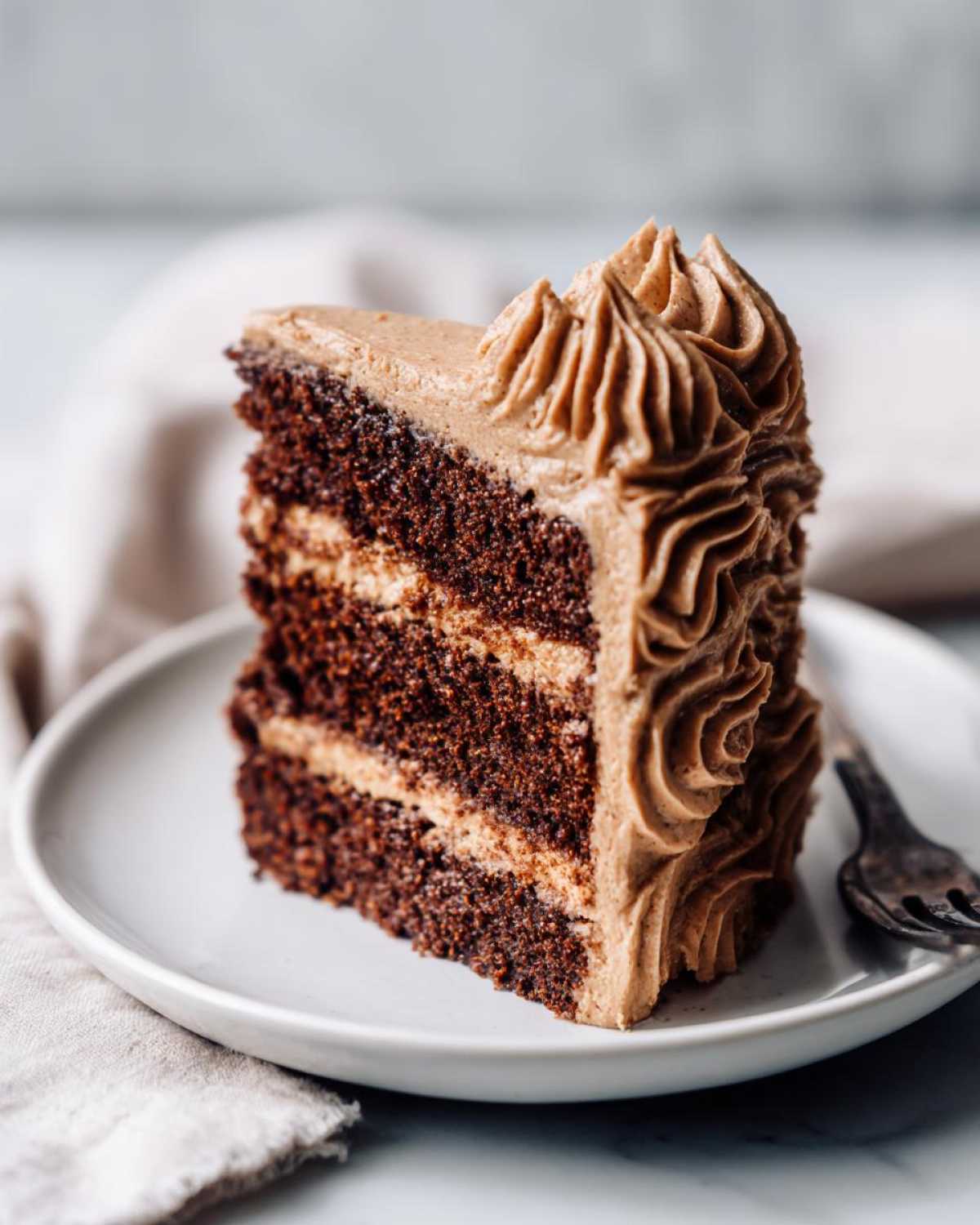 A tall slice of dark chocolate gingerbread latte cake with layers of light brown frosting, served on a white plate.