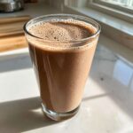 A tall glass filled with freshly made chocolate milk, showing a frothy top, sitting on a sunny white counter.