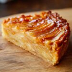 A close-up, cross-section view of a rich, caramelized slice of apple tart resting on a wooden board.