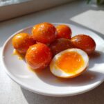 A close-up of several glossy, orange marinated eggs on a white plate, one cut in half showing a jammy yolk.
