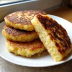 A stack of freshly cooked, golden brown irish potato farls on a white plate, with one piece cut to show the soft interior.