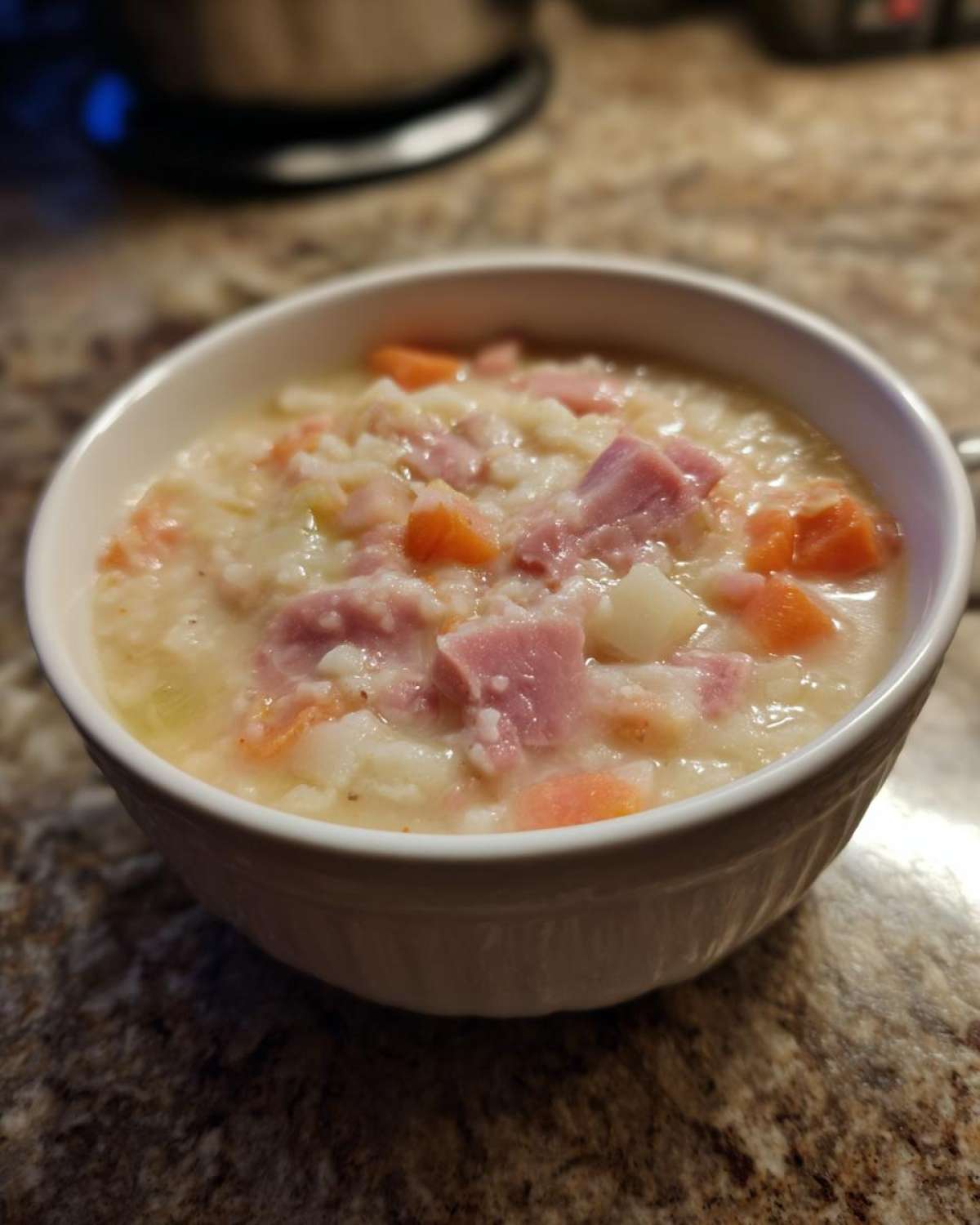 A close-up of a creamy, hearty bean soup with chunks of ham, carrots, and potatoes served in a white bowl.